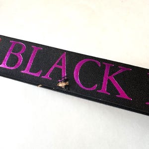 K&ouml;nnte beinhalten: Nahaufnahme eines schwarzen Bandes mit dem Wort "BLACK" in leuchtend pinkfarbenen Metallic-Lettern. Das Band hat einen kleinen Riss in der N&auml;he der Mitte. Der Hintergrund ist wei&szlig;.