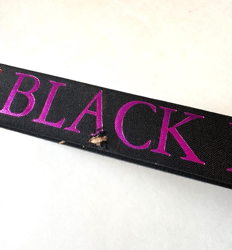 K&ouml;nnte beinhalten: Nahaufnahme eines schwarzen Bandes mit dem Wort "BLACK" in leuchtend pinkfarbenen Metallic-Lettern. Das Band hat einen kleinen Riss in der N&auml;he der Mitte. Der Hintergrund ist wei&szlig;.