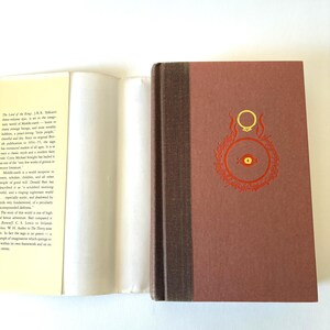 May include: A hardcover book with a brown cover featuring a gold ring design and red accents. The spine is a darker brown. The book is open, revealing white pages and text. The book is a classic fantasy novel.