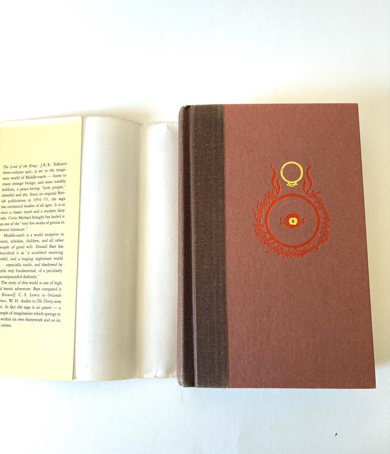 May include: A hardcover book with a brown cover featuring a gold ring design and red accents. The spine is a darker brown. The book is open, revealing white pages and text. The book is a classic fantasy novel.