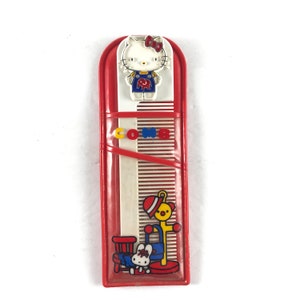 May include: Red plastic comb case with a Hello Kitty character on the front. The case has a white comb inside with a Hello Kitty character on the bottom.