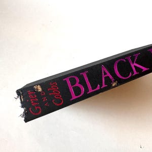 K&ouml;nnte beinhalten: Nahaufnahme eines Buchr&uuml;ckens mit dem Titel "BLACK" in leuchtend pinkfarbenen Buchstaben. Der Name des Autors, "Grier and Cobbs", ist rot gedruckt. Der Buchdeckel ist schwarz, und die Kanten weisen Abnutzungserscheinungen auf.