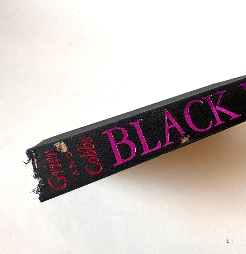 K&ouml;nnte beinhalten: Nahaufnahme eines Buchr&uuml;ckens mit dem Titel "BLACK" in leuchtend pinkfarbenen Buchstaben. Der Name des Autors, "Grier and Cobbs", ist rot gedruckt. Der Buchdeckel ist schwarz, und die Kanten weisen Abnutzungserscheinungen auf.