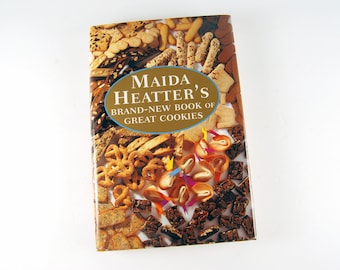 May include: A cookbook titled "Maida Heatter's Brand-New Book of Great Cookies" with a cover featuring an assortment of cookies. The cover has a gold oval with the title and author's name. Various cookies are arranged around the title.