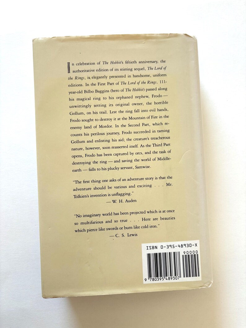 May include: A book cover for "The Lord of the Rings" by J.R.R. Tolkien. The back cover is a light beige color with black text describing the story and quotes from W.H. Auden and C.S. Lewis. The ISBN is visible.