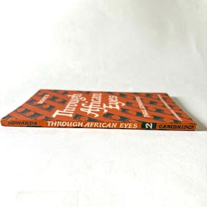 Pu&ograve; includere: Un libro vintage intitolato "Through African Eyes" di Paul Strand, con un motivo geometrico rosso e nero sulla copertina. Pubblicato da Cambridge, con il numero 2 sul dorso.