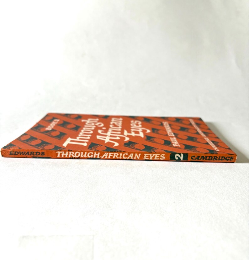 Pu&ograve; includere: Un libro vintage intitolato "Through African Eyes" di Paul Strand, con un motivo geometrico rosso e nero sulla copertina. Pubblicato da Cambridge, con il numero 2 sul dorso.