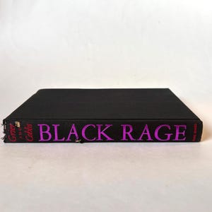 K&ouml;nnte beinhalten: Ein schwarzes Hardcover-Buch mit dem Titel "BLACK RAGE" in leuchtend rosa Buchstaben. Der Buchr&uuml;cken zeigt auch die Namen der Autoren, "Grier and Cobbs", in Rosa. Das Buch ist vor einem wei&szlig;en Hintergrund platziert.