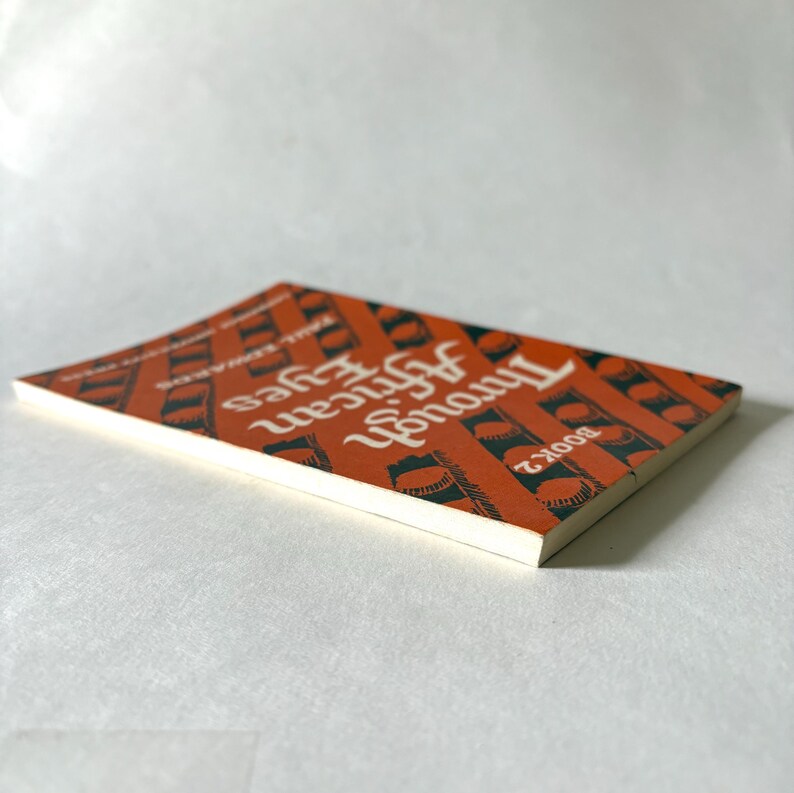 Pu&ograve; includere: Un libro tascabile intitolato "Through African Eyes Book 2" con una copertina arancione con motivi geometrici neri e testo bianco. Il dorso del libro &egrave; visibile e le pagine sono bianche.