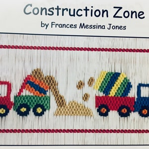 May include: Embroidered artwork titled "Construction Zone" by Frances Messina Jones. The design features colorful construction vehicles: a red truck, a green excavator, a red truck with a striped mixer, and a blue truck. The background is white with red trim.