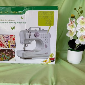 May include: A white and gray mini sewing machine with the text "Mini Multifunctional Household Sewing Machine" on the box. The box also displays fabric samples. A white potted orchid with white and pink flowers is to the right.