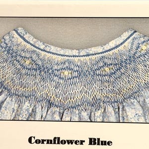 May include: A close-up of a blue and white floral fabric with intricate smocking detail. The neckline is adorned with delicate stitching and a scalloped edge. The text "Cornflower Blue" is at the bottom of the image.