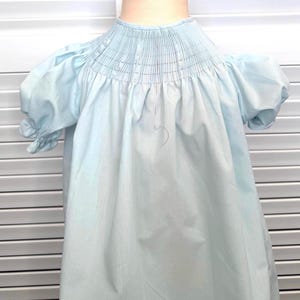 May include: A light blue dress featuring a smocked yoke with delicate vertical stitching, short puff sleeves with gathered cuffs, and a loose, flowing skirt. The garment is displayed on a mannequin, showcasing the detailed pleating at the neckline.