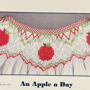 May include: White fabric with red apple embroidery, green accents, and a red and white checkered trim. The text "An Apple a Day" is at the bottom. The design is a close-up of a garment.