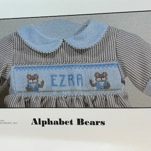 May include: A baby's romper with a blue and white striped pattern, featuring a light blue collar and cuffs. The front panel has a cross-stitched design with the name "EZRA" and two bear illustrations. The text "Alphabet Bears" is below the romper.
