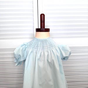 May include: A light blue baby dress with a smocked bodice and short, puffed sleeves. The dress is displayed on a wooden dress form against a white shutter background. The dress has a simple, classic design.