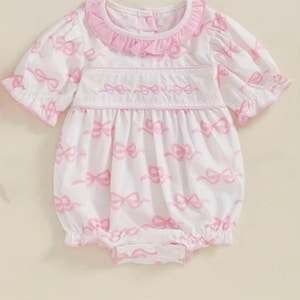 May include: White baby romper with a pink bow pattern. The romper features a ruffled collar and sleeves, with embroidered bows across the chest. The garment is designed with a bubble silhouette and a snap closure.