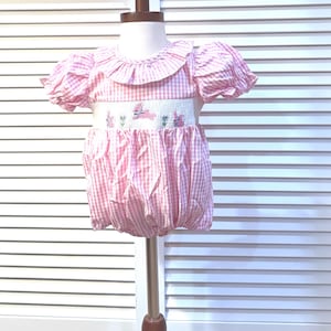 May include: Pink and white gingham baby romper with a ruffled collar and puffed sleeves. The romper features a white smocked panel with embroidered bunnies and flowers. The bottom is gathered, creating a bubble shape.