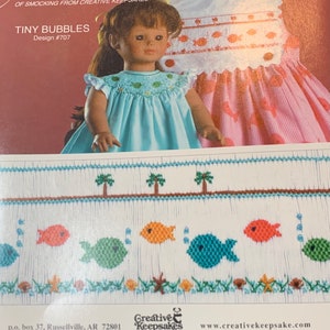 May include: A doll wearing a light blue smocked dress with a ruffled collar. The dress features embroidered fish and a matching design on the bodice. The image also shows a pattern with embroidered fish, palm trees, and sea elements.