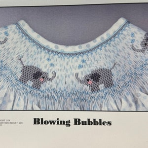 May include: A light blue baby dress with a smocked bodice and elephant embroidery. The dress features a pattern of small blue polka dots and the text "Blowing Bubbles".