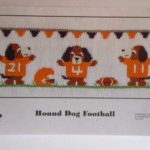May include: Cross-stitch design featuring three cartoon hound dogs in orange football jerseys with numbers, a football, and a helmet. The text "Hound Dog Football" is below the image, with a banner of orange and brown pennants above.