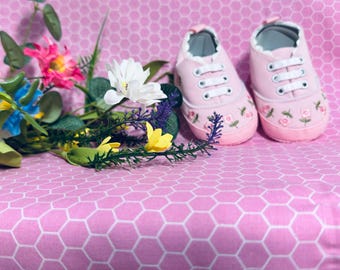 Op de afbeelding: Een paar roze babysneakers met witte veters en bloemenborduurwerk. De schoenen staan op een roze zeshoekig patroon, naast een boeket kleurrijke kunstbloemen.
