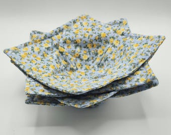 May include: A stack of four fabric bowl cozies. Each cozy is light blue with a pattern of small yellow flowers and green leaves. The cozies are square with folded corners, creating a bowl shape.