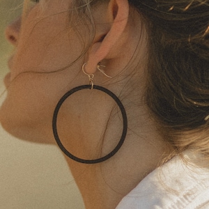 May include: A close-up shot of a person wearing large, black hoop earrings. The earrings are circular and hang from gold-colored hooks. The person is wearing a white shirt, and their hair is pulled back.