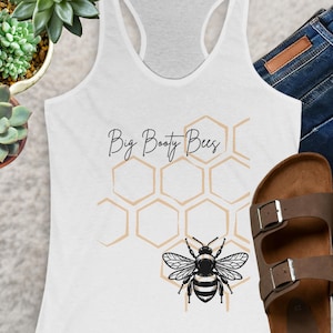 May include: White vest top featuring the text "Big Booty Bees". The design includes a honeycomb pattern and a bee illustration. The top is styled with blue denim jeans and brown sandals.