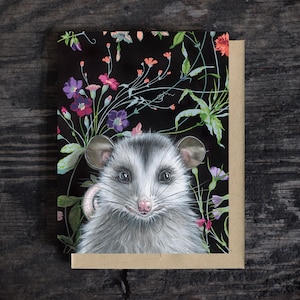 Peut inclure: Une carte de vœux rectangulaire présentant une illustration détaillée d'un opossum gris et blanc centré sur un fond noir rempli de fleurs sauvages colorées dans des tons de rose, violet et vert, accompagnée d'une enveloppe beige unie.