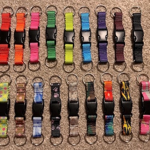 May include: Twenty keychains with different colored and patterned straps and silver key rings. The straps are made of a woven material and have a black plastic buckle.