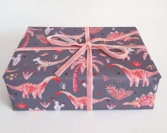 May include: A gray gift box with a pink ribbon. The box is wrapped in wrapping paper with a pattern of pink dinosaurs and palm trees.