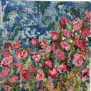 May include: A square textile wall art piece featuring a dense, textured punch needle or rug hooking design. The composition displays a vibrant garden scene with clusters of pink and red flowers set against a background of blue and green foliage.