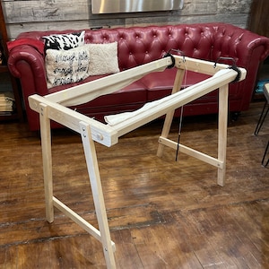 May include: A wooden embroidery hoop stand with a white fabric stretched across the hoop. The stand is made of natural wood and has a simple design. The stand is in a room with a red leather sofa and a pillow with the text "enough faith enough love enough to be enough".