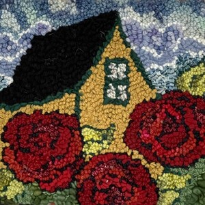 May include: A rug hooking artwork featuring a yellow house with a black roof, surrounded by red flowers and green foliage. The background is a blue sky with white clouds.