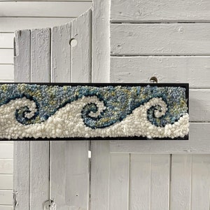 May include: A framed wall hanging with a blue and white wave design. The waves are made of yarn and are textured. The frame is black.