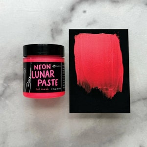 Puede incluir: Un pequeño tarro de pasta lunar neón rosa intenso, etiquetado como "Neon Lunar Paste" con el nombre de la marca "Ranger" y el texto "hot mess" debajo. El tarro está sobre una superficie de mármol blanco.