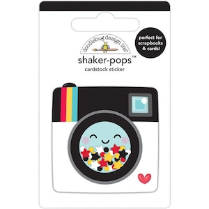 May include: A cardstock sticker of a black and white camera with a rainbow stripe on the side. The camera lens is a shaker-pops design with a smiling face and colorful stars inside. The text "shaker-pops" and "cardstock sticker" is printed on the sticker.