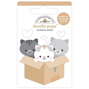 May include: Cardstock sticker featuring three cartoon cats peeking out of a cardboard box. The cats are gray and white with pink cheeks. The packaging includes the text "doodle-pops" and "perfect for scrapbooks & cards!"