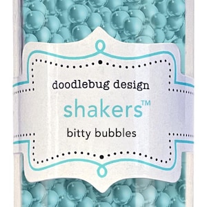 May include: A clear glass jar filled with light blue, spherical beads. The jar has a silver lid and a label that reads "doodlebug design shakers bitty bubbles."