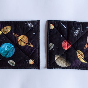 May include: Two black quilted pot holders with a space theme. The pot holders feature a black background with colorful planets, stars, and a comet.
