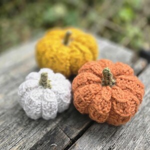 May include: Three small, round, crocheted pumpkins in white, mustard yellow, and burnt orange. Each pumpkin features a textured, ribbed surface and a short, green, crocheted stem at the top centre. The pumpkins are arranged in a cluster.