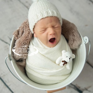 May include: A newborn baby wearing a white knit hat with a white pom pom, wrapped in a white blanket, and holding a white teddy bear. The baby is in a white bucket with a brown furry blanket in the background. The image includes the text "Santana Nicole Photography".