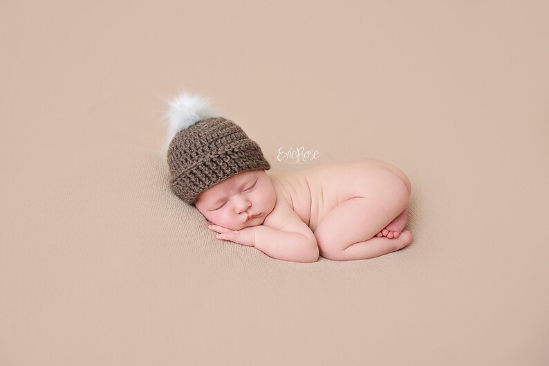 A newborn baby wearing a brown crocheted hat with a white pom-pom, sleeping on a light brown background. *created following a crochet pattern from Lily B's, also known as lilybphotoprops, to make a crocheted Pumpkin Hat - this version is the year round hat minus the pumpkin leaf.