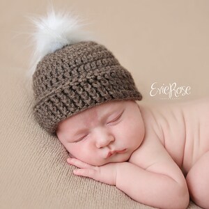 Puede incluir: Un gorro de ganchillo marr&oacute;n con un borde acanalado y un pomp&oacute;n blanco esponjoso en la parte superior. El gorro est&aacute; construido con un patr&oacute;n de hilo texturizado y descansa sobre una superficie. El texto EvieRose Photography es visible.