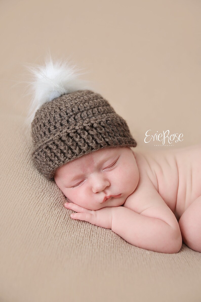 Puede incluir: Un gorro de ganchillo marr&oacute;n con un borde acanalado y un pomp&oacute;n blanco esponjoso en la parte superior. El gorro est&aacute; construido con un patr&oacute;n de hilo texturizado y descansa sobre una superficie. El texto EvieRose Photography es visible.