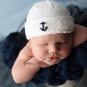 May include: A white knitted beanie with a folded brim featuring a small, dark blue embroidered anchor emblem on the front. The hat is worn by a sleeping infant nestled within a dark navy blue, chunky knit fabric wrap with a soft texture.