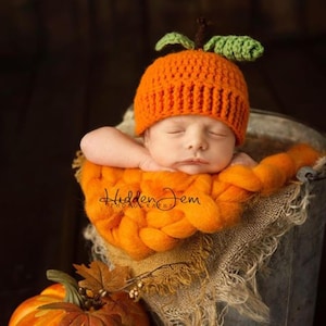 The image shows a sleeping newborn baby, posed in a bucket against a dark background, atop a thick, orange knitted blanket. Baby is wearing a crocheted pumpkin hat created using a crochet pattern from Lily B's.