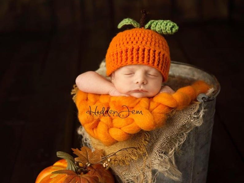 The image shows a sleeping newborn baby, posed in a bucket against a dark background, atop a thick, orange knitted blanket. Baby is wearing a crocheted pumpkin hat created using a crochet pattern from Lily B's.