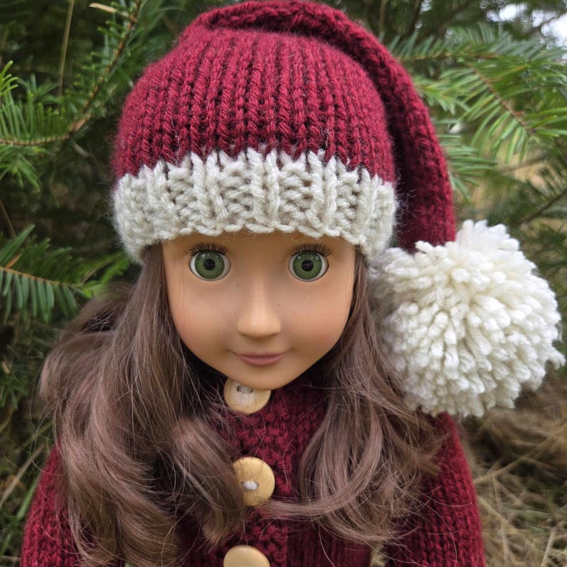 Pu&ograve; includere: Una bambola con lunghi capelli castani indossa un berretto in maglia rosso scuro con un bordo a coste color crema e un grande pompon color crema sulla punta. La bambola indossa un cardigan in maglia rosso scuro abbinato con tre bottoni in legno.